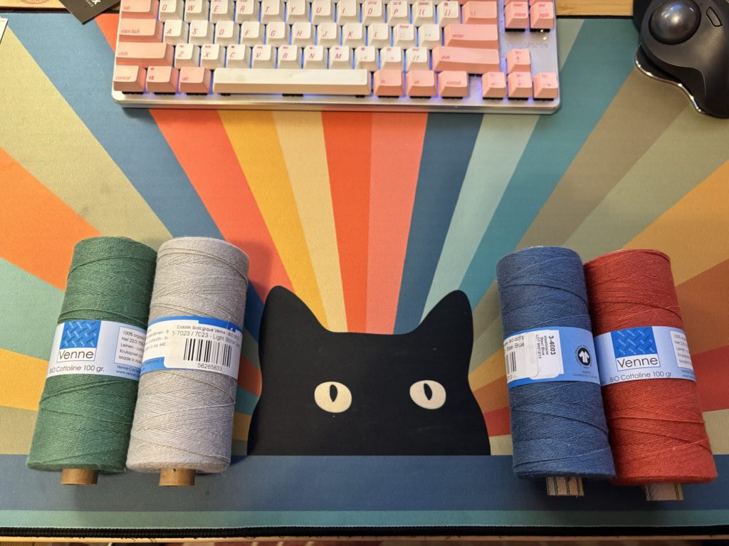 Four spools of cottolin yarn, on my desk mat, which features black cat peeking over the edge, with rainbow beaming behind it.