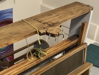 The top of my table loom, showing four small holes and improvised hooks jutting out of a semicircular hub of wood. Yellow yarn connected to the shafts dangles.